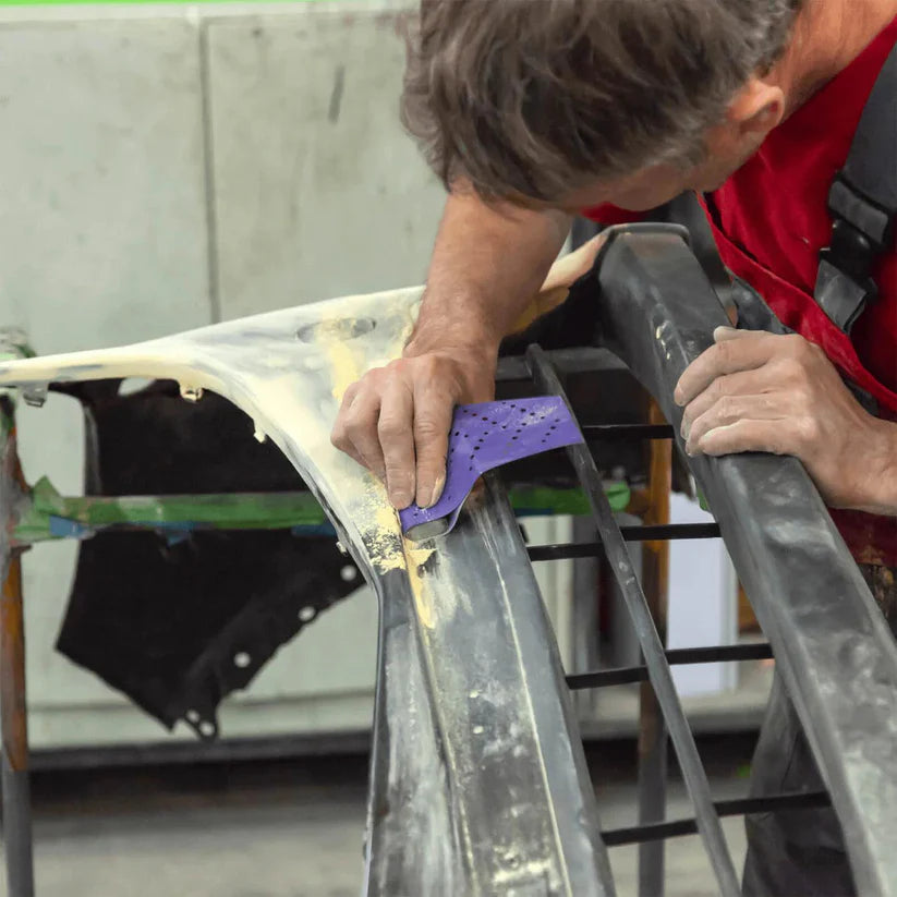 Person sanding on a car panel with sanding tools and sanding sheet, wearing a red shirt and gray pants.