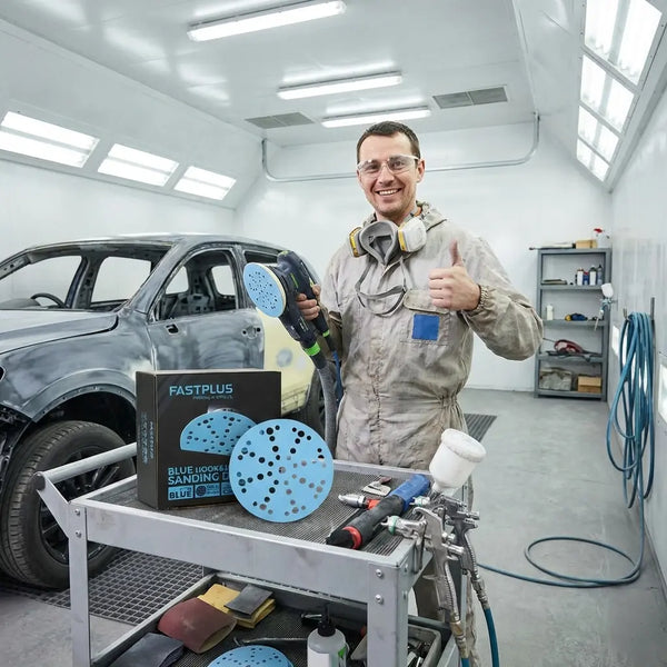 Person in a paint booth with automotive tools and equipment, holding a box of sanding discs labeled 'Fastplus'.